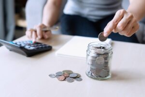 יד מניחה מטבע בצנצנת חיסכון זכוכית ליד מחשבון ומחברת על שולחן